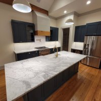 Spacious open-concept kitchen with dark navy cabinets, white shiplap hood, marble island countertops, stainless steel refrigerator, pendant lights, and wooden beams