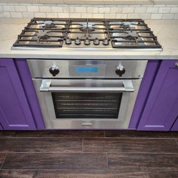 A kitchen with a stainless steel gas stove top and oven, white subway tile backsplash, and purple cabinetry.