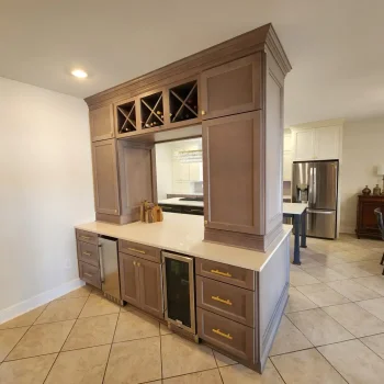 Peninsula Kitchen Cabinetry, light brown cabinetry with gold hardware and white quartz countertops wine cooler and ice maker. Wine racks with wine bottles in them