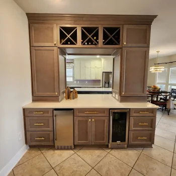 Peninsula Kitchen Cabinetry, light brown cabinetry with gold hardware and white quartz countertops wine cooler and ice maker. Wine racks with wine bottles in them front shot