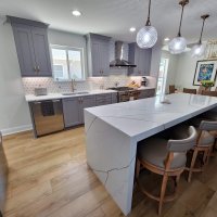 Blue gray kitchen cabinetry with white quartz countertops that has dark veining, cabinetry has gold hardware tile backsplash has a sunburst pattern. Kitchen Island has bookmatched laminated 3 inch counter in quartz