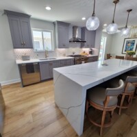 Blue gray kitchen cabinetry with white quartz countertops that has dark veining, cabinetry has gold hardware tile backsplash has a sunburst pattern. Kitchen Island has bookmatched laminated 3 inch counter in quartz