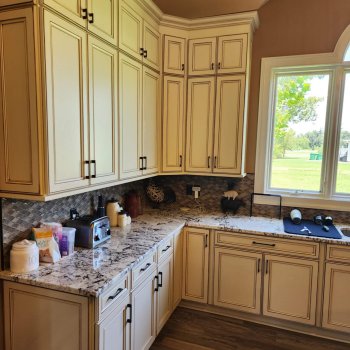 A corner of the kitchen with beige cabinets, a marble or granite countertop, and a patterned tile backsplash. There are various kitchen items and decor on the countertop.