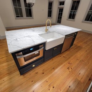 Timeless luxury with Algiers Point marble countertops in Orleans Parish. This overall view highlights an expansive kitchen island topped with premium, large-format Carrara marble. The stone is meticulously fabricated to house a central white porcelain farm-style sink, creating a classic contrast against the custom blue island cabinetry. Algiers Point marble countertops featuring a large Carrara marble kitchen island with an undermount apron-front farmhouse sink over blue cabinetry in New Orleans.