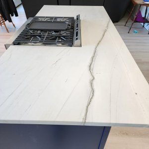 Transform your home with luxury Luling quartzite countertops in St. Charles Parish. This stunning island highlights Mont Blanc Quartzite, displaying a bright cream background split by a prominent, linear gray vein tracking across the stone. A professional gas cooktop is drop-in mounted directly into the center of the slab. Luling quartzite countertops showing a light cream Mont Blanc Quartzite island with a striking linear gray vein and a built-in gas cooktop in St. Charles Parish.